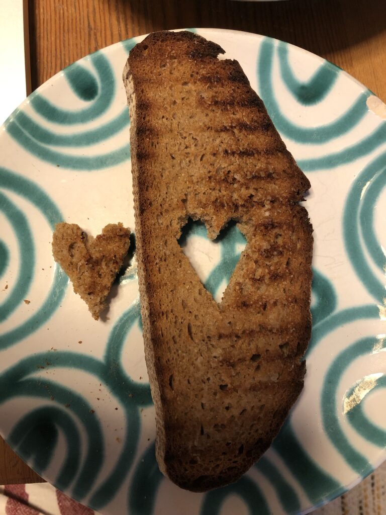 Ein Brot, bei dem in der Mitte ein Herz ausgeschnitten ist auf einem Teller. Das Herz liegt daneben. 
