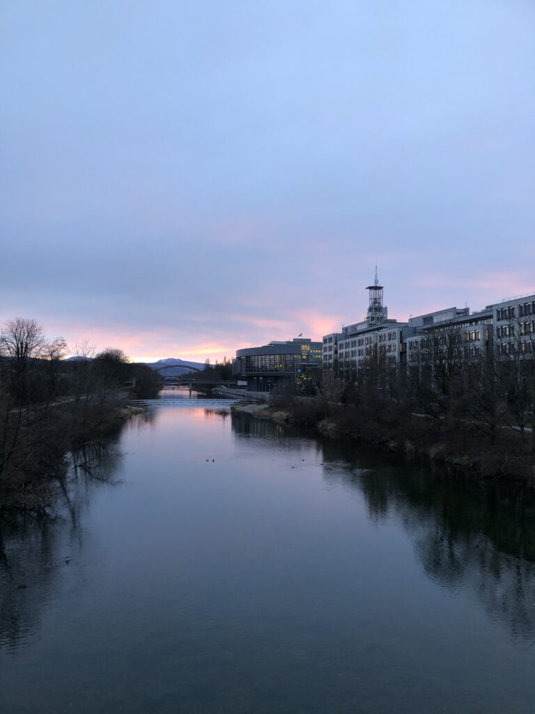 Im Vordergrund ein Fluss, rechts Häuser und links ein kleiner Weg am Flussufer. Im Hintergrund der Sonnenuntergang mit lila, orangefarbenem Himmel.