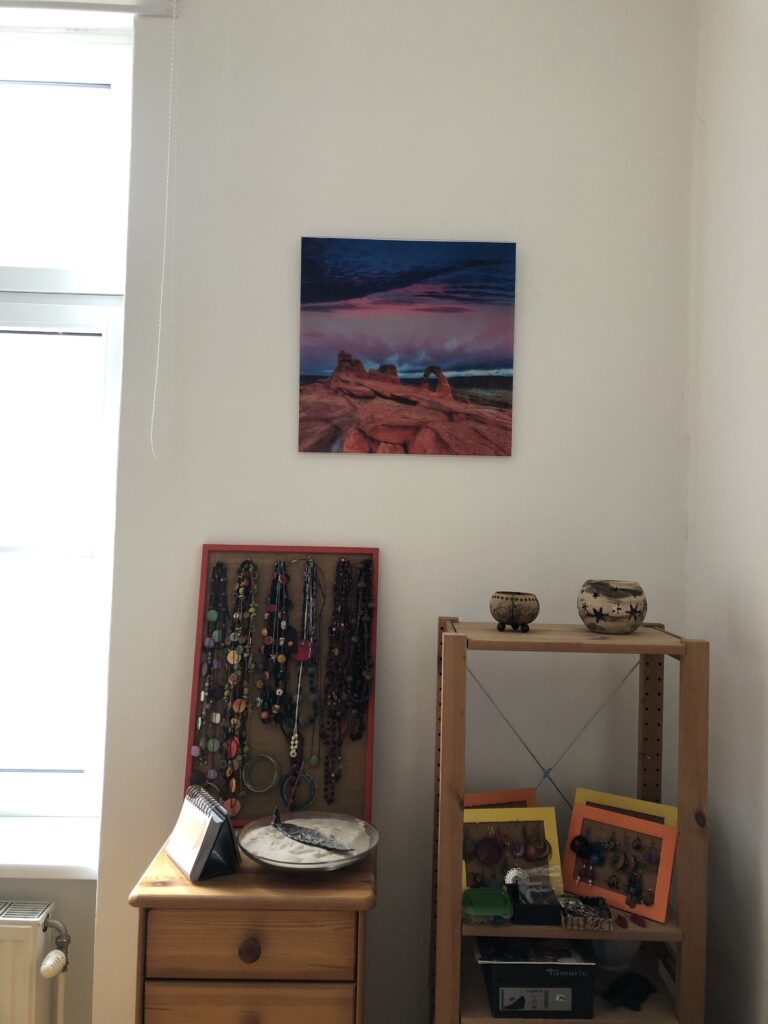 Ein Foto einer Wand in meiner Wohnung, an der jetzt ein Glasbild mit einer Steinformation am Meer hängt. Darunter Schmuck in Bilderrahmen aufgehängt auf Regalen stehend. 