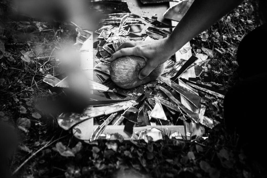 schwarz-weiß Bild, wie jemand mit einem großen Stein einen Spiegel zertrümmert, der in 1000 Scherben bricht