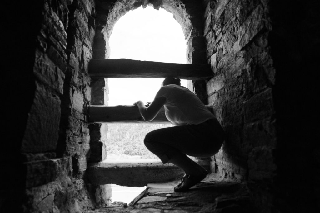schwarzweiß, Frau, die in einem Steinturm durch drei dicke Balken nach draußen schaut - ein Sinnbild für das Gefangensein im transgenerationalen Trauma
