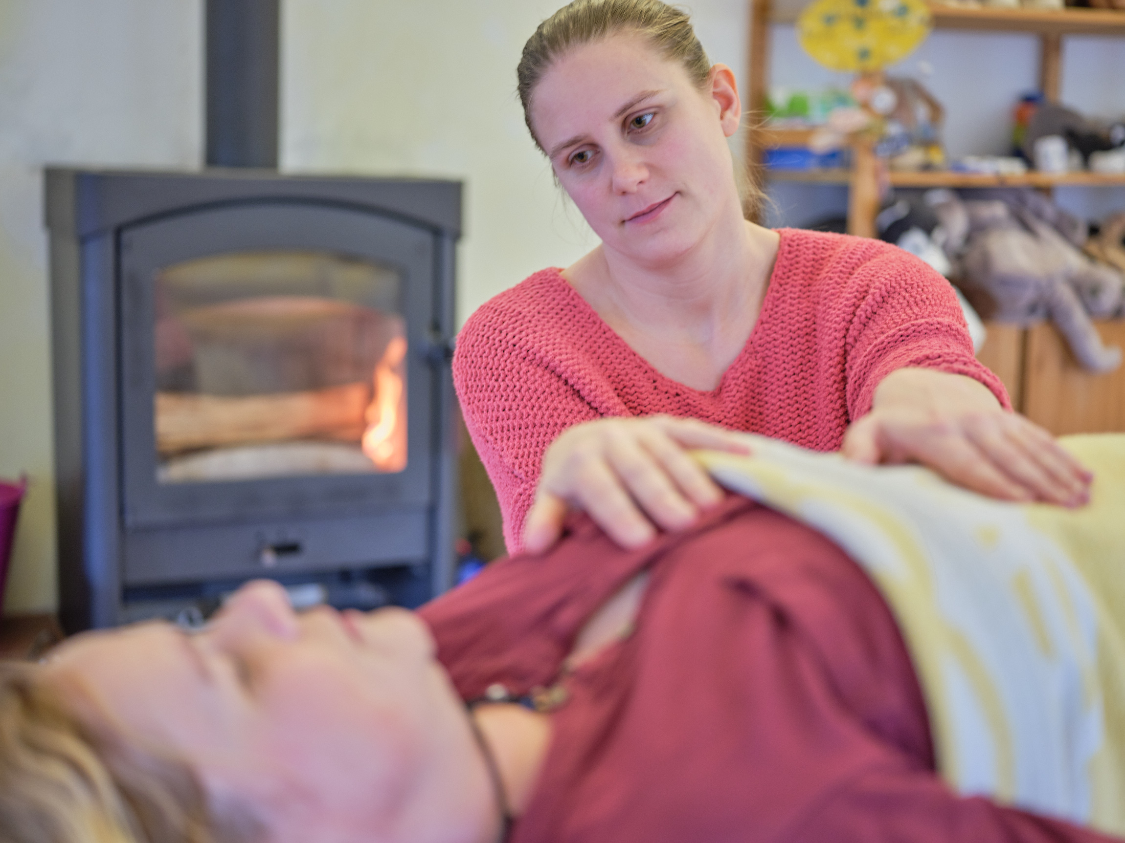 Eine Person im rosa Pullover legt sanft ihre Hände auf die Brust einer anderen Person, die unter einer Decke liegt. Im Hintergrund glüht ein brennendes Kaminfeuer, während sie sich in dieser warmen und ruhigen Umgebung auf Körpersignale deuten konzentriert.