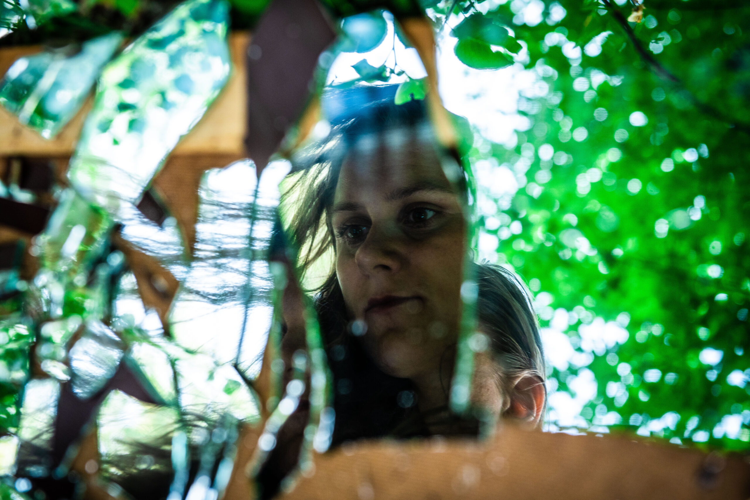 Transgenerationale Weitergabe_3 Eine Frau schaut in einen zerbrochenen Spiegel, der im Gras liegt. Ein Sinnbild für unerforschte transgenerationale Weitergabe, die oft ein Gefühl von Trennung, Ausweglosigkeit und Ratlosigkeit mit sich bringt.