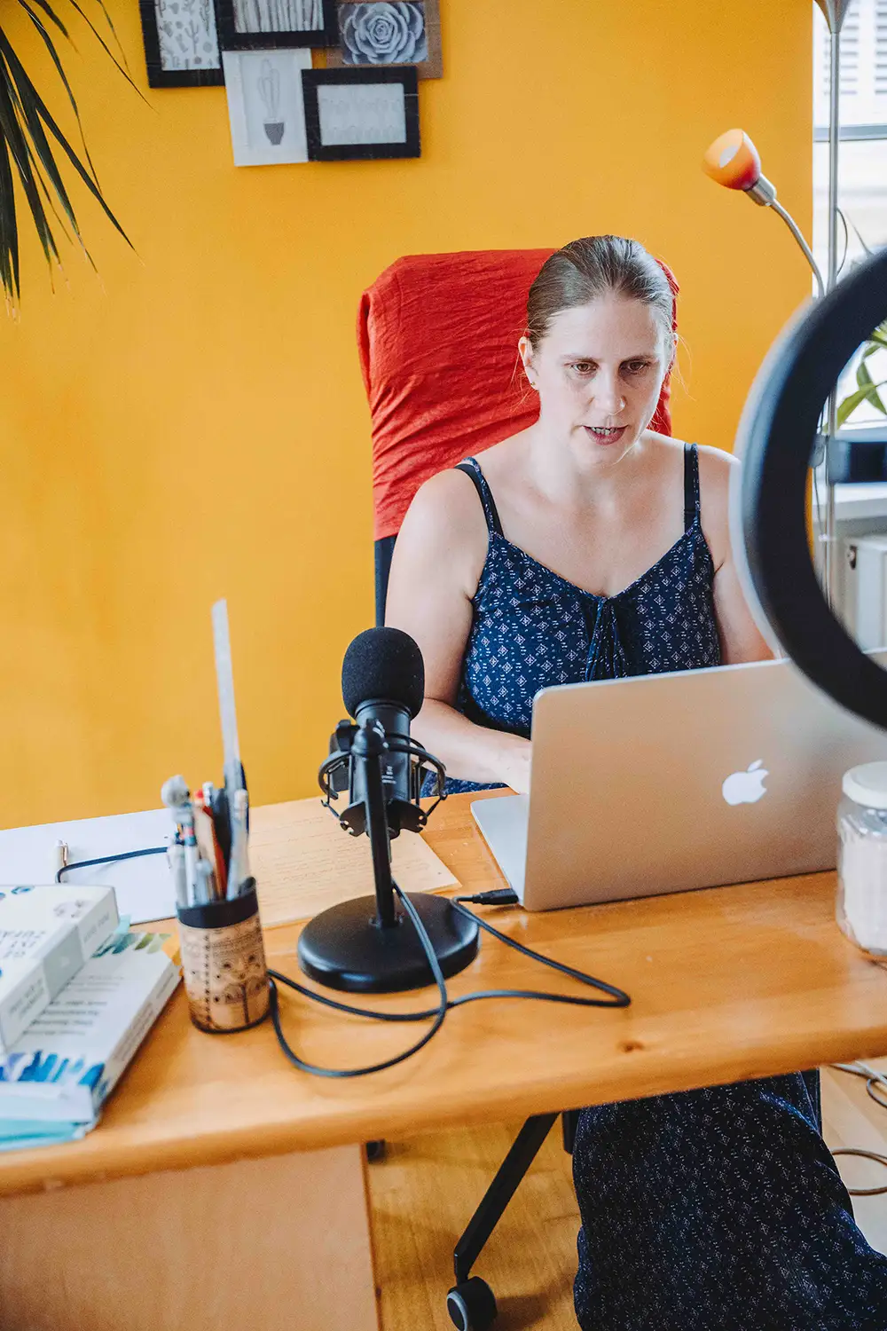 Generose sitzt mit einem Laptop und einem Mikrofon an einem Holzschreibtisch vor einer orangefarbenen Wand mit gerahmter Kunst.