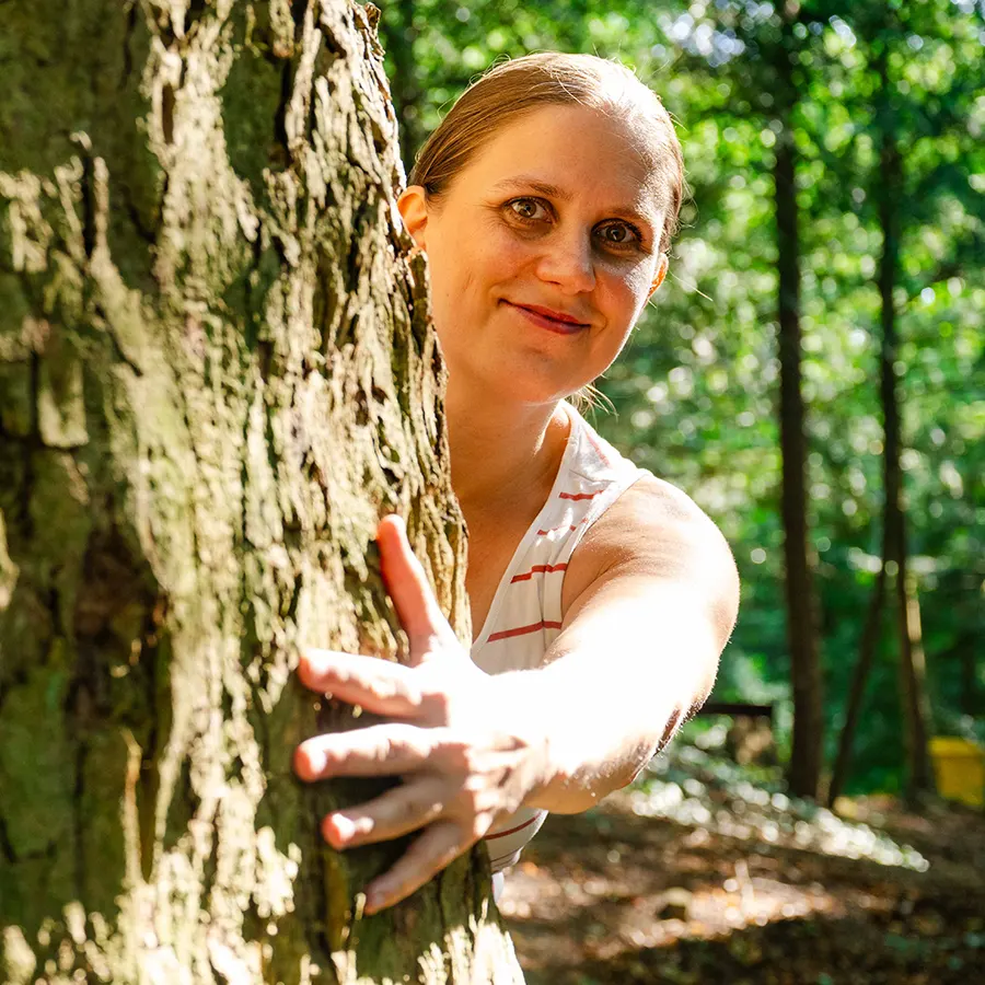 Eine Frau lächelt, während sie in einem sonnendurchfluteten Wald einen Baum umarmt und ihre Hand um den Stamm legt. Geflecktes Sonnenlicht fällt durch die grünen Blätter im Hintergrund.