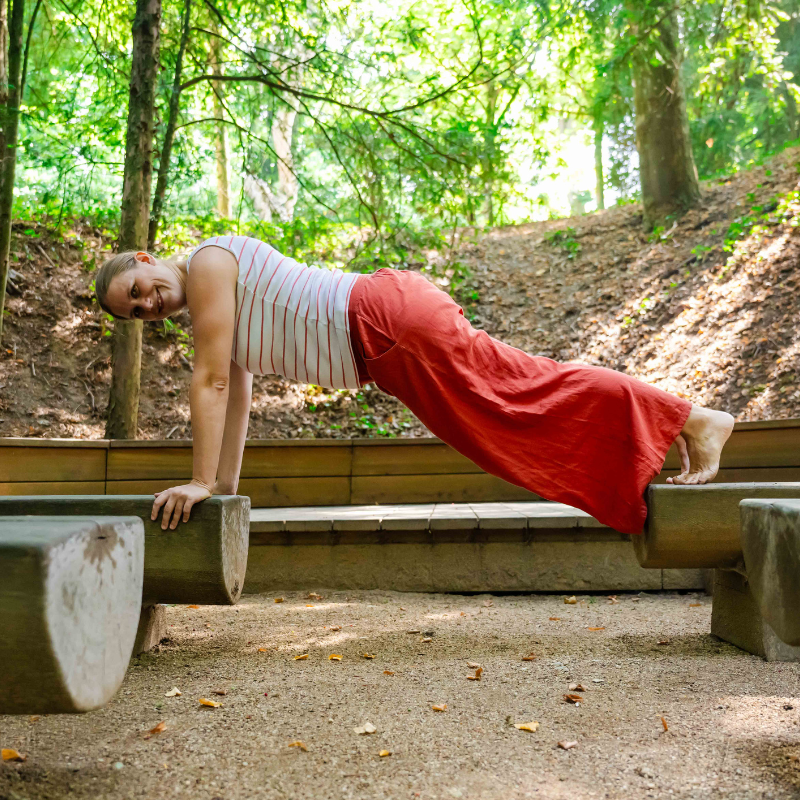 Eine Frau in einem weiß gestreiften Oberteil und einer roten Hose balanciert in einer Plankenposition mit ihren Händen auf einer Holzbank und ihren Füßen auf einer anderen, im Freien in einem Waldstück, und lächelt in die Kamera. Symbolisiert die NEIN-Challenge.