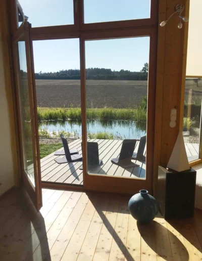 Sonnenlicht fällt durch große Glastüren in einen Raum mit Holzboden und führt zu einer Terrasse mit Stühlen und Blick auf einen Teich, eine Wiese und Bäume in der Ferne. Im Inneren stehen eine blaue Vase und ein Dekorationsobjekt.
