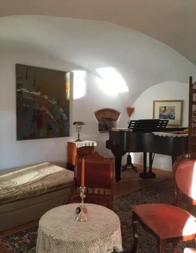 Ein sonnendurchfluteter Raum mit einem Flügel, einer Chaiselongue, roten antiken Stühlen, einem runden Tisch mit Spitzendecke, einem gemusterten Teppich und abstrakter Kunst an der Wand. Der Raum hat gewölbte weiße Decken und ein klassisches Dekor.