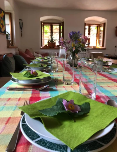 Ein gedeckter Tisch mit grünen Servietten, Blättern und rosa Blumen auf jedem Teller, ordentlich platzierten Gläsern und Besteck und einer karierten Tischdecke. Sonnenlicht fällt durch die offenen Fenster in den gemütlichen Raum.