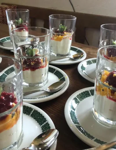 Mehrere Gläser mit cremigem Dessert, garniert mit Fruchtsauce und Minzblättern, sind auf Tellern mit Löffeln auf einem Holztisch angeordnet.