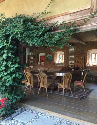 Ein rustikaler Essbereich mit Korbstühlen um einen Holztisch, Backsteinwänden und Kunstwerken. Üppige grüne Ranken hängen von einem Holzbalken am Eingang, und draußen ist der Boden mit Kieselsteinen bedeckt. Warmes Sonnenlicht fällt durch kleine Fenster.
