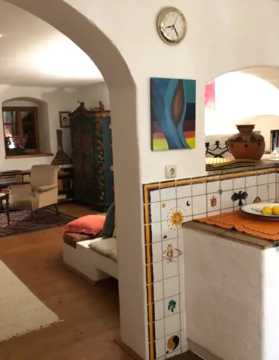 Ein gemütlicher Wohnraum mit weißen Wänden, gewölbten Türen, Holzböden und farbenfrohen Fliesenakzenten. Der Raum verfügt über bequeme Sitzgelegenheiten, einen Teppich, dekorative Möbel, Kunstwerke und eine kleine Uhr an der Wand.