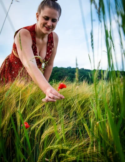 Eine Frau in einem rot gemusterten Kleid lächelt, während sie sanft eine rote Mohnblume auf einem Feld mit hohem grünem Gras berührt, im Hintergrund Bäume und ein blauer Himmel.