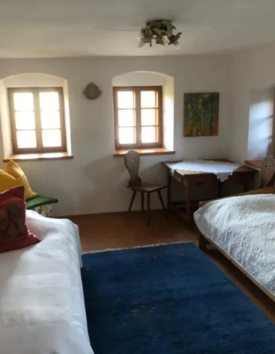 Gemütliches Schlafzimmer mit zwei Einzelbetten, bunten Kissen, einem blauen Teppich, Holzmöbeln und Sonnenlicht, das durch drei Fenster fällt. Gemälde und ein kleiner Tisch verleihen dem weiß getünchten Zimmer Charme.