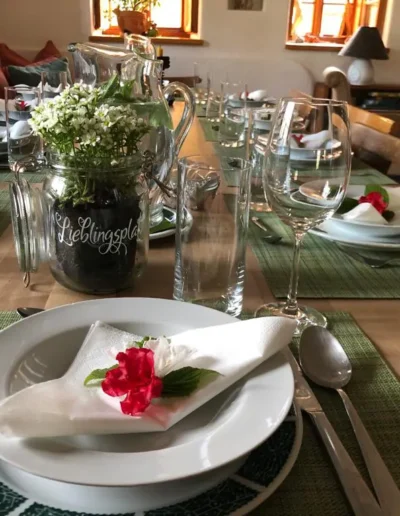 Ein Esstisch ist mit weißen Tellern, Gläsern, Besteck und grünen Tischsets gedeckt. Auf jedem Teller liegen Servietten mit roten Blumen. Ein Glasgefäß mit weißen Blumen dient als Tafelaufsatz. Warmes Sonnenlicht fällt durch die Fenster.