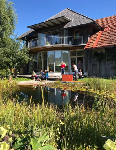 An einem sonnigen Tag sitzt und steht eine Gruppe von Menschen an einem kleinen Teich vor einem modernen Holzhaus mit großen Fenstern, einem Balkon und einem schrägen Dach. Üppiges Grün und Blumen umgeben den Teich.