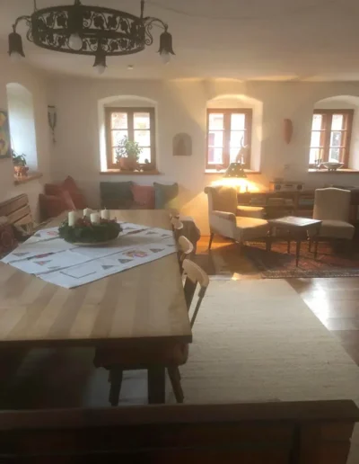 Ein gemütliches, sonnendurchflutetes Esszimmer mit Holztisch und -stühlen, Kerzenhalter, Papier auf dem Tisch, gepolsterten Bänken, Sesseln, einem Teppich und mehreren Fenstern, die natürliches Licht hereinlassen. Warmes, einladendes Dekor und sanfte Beleuchtung.