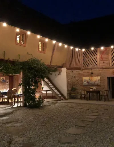 Ein gemütlicher Innenhof bei Nacht, beleuchtet von Lichterketten, mit rustikalen Backsteinmauern, Holzbalken, Pflanzen, Sitzgelegenheiten im Freien und Kunstwerken an den Wänden. Ein Steinweg führt durch den Kieshof.