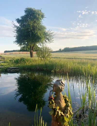 Eine friedliche ländliche Landschaft mit einem Teich, in dem sich ein großer Baum und der blaue Himmel spiegeln. Hohes Gras säumt den Wasserrand, und im Vordergrund steht eine kleine Statue mit einem Blumenkranz. Im Hintergrund erstrecken sich Felder.