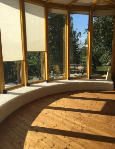 Ein sonnendurchfluteter Raum mit großen, hohen Fenstern, teilweise herunterziehbaren Jalousien, geschwungenen weißen Sitzgelegenheiten und Holzboden. Durch das Glas sind Bäume und Grünflächen draußen sichtbar.