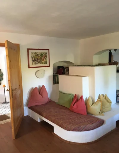 Eine gemütliche Einbaunische mit gepolsterter Bank, bunten Kissen, einem kleinen Bücherregal und gerahmter Kunst an der Wand. Der Bereich ist in warmen Tönen gehalten und verfügt über eine Holztür, die zu einem angrenzenden Raum führt.