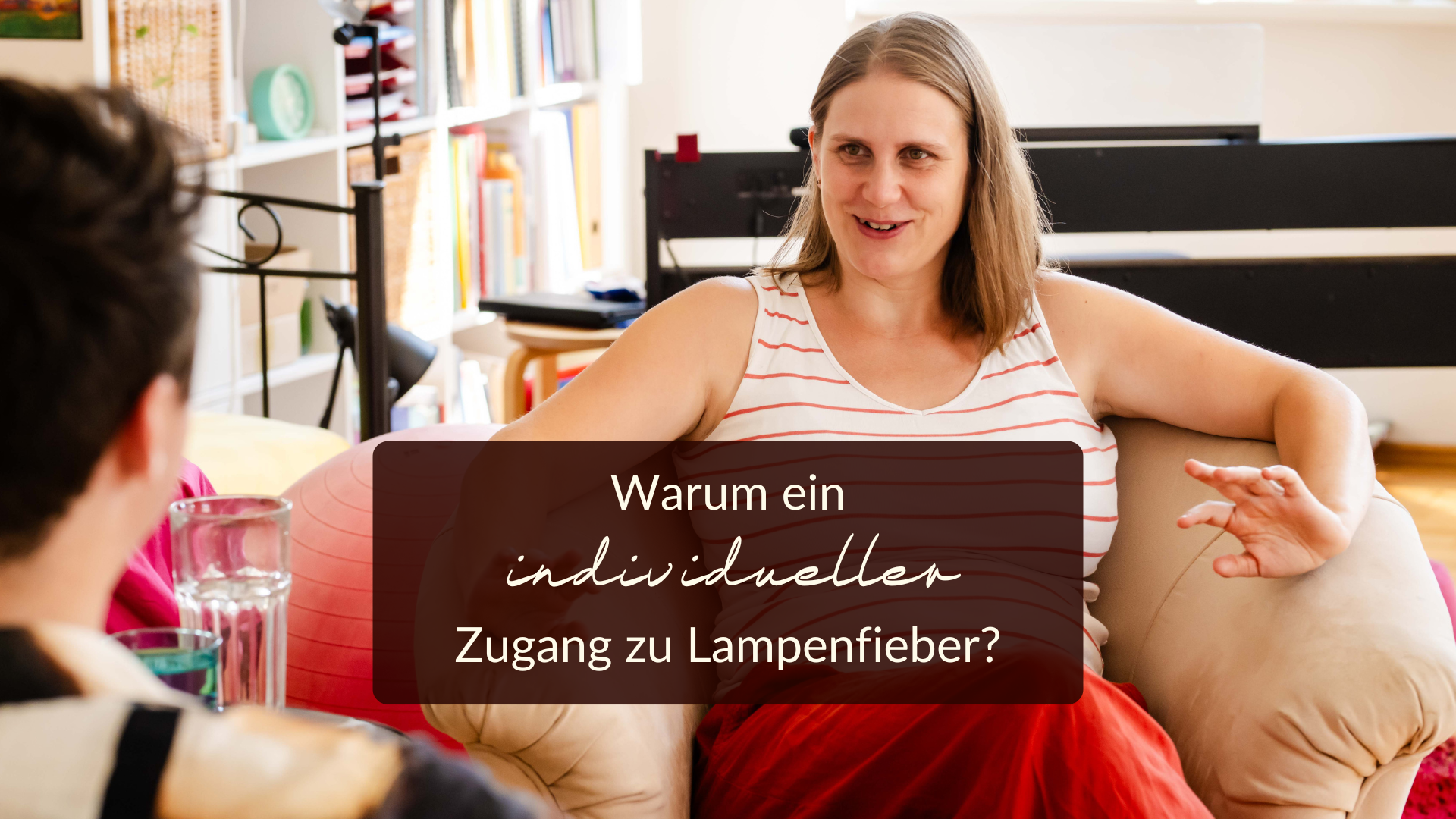 individueller Zugang Lampenfieber Eine Frau in einem roten Rock sitzt auf einem beigen Stuhl und spricht mit jemandem aus dem Off. Der deutsche Text auf dem Bild lautet: "Warum ein individueller Zugang zu Lampenfieber?" Im Hintergrund sind Bücherregale und helles Dekor zu sehen.