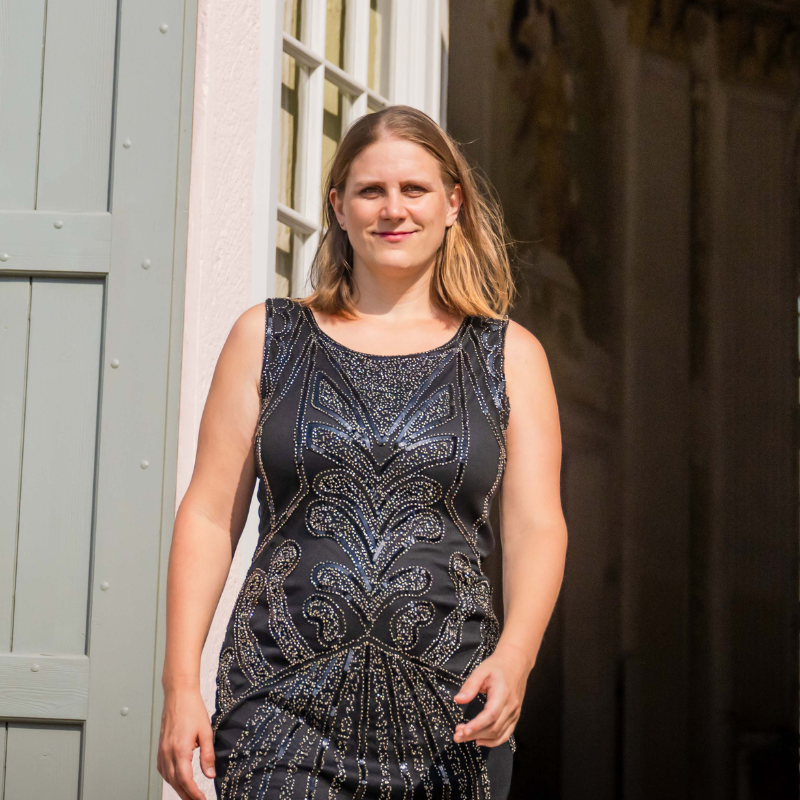 Eine Frau in einem paillettenbestickten Kleid schaut selbstbewusst in die Kamera. Im Hintergrund eine graue Tür. Die Frau ist gerade mitten im Gehen. Symbol für einen selbstbewussten Auftritt - Höchstleistung trotz Lampenfieber.