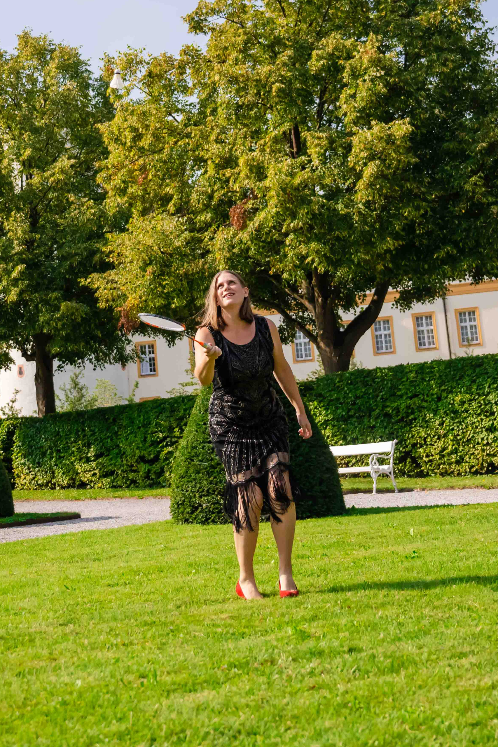 Souverän & kraftvoll auf der Bühne 2 Eine Frau spielt Badminton in einem Paillettenkleid mitten auf der Wiese. Im Hintergrund ist ein Haus zu sehen und zwei Bäume, außerdem eine weiße Parkbank. Das Bild ist ein Symbol für spielerische Qualität auf der Bühne.