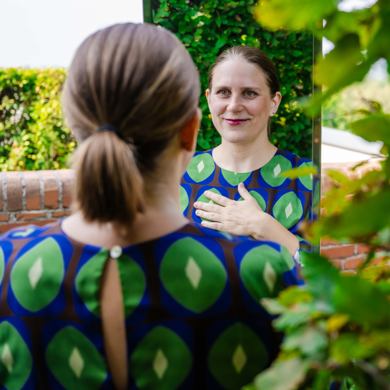 Eine Frau, in einem grün-blauen Kleid, von hinten fotografiert. Sie steht vor dem Spiegel und sieht sich selbst in die Augen, dabei hat sie die rechte Hand auf ihren Brustkorb gelegt. Ein Sinnbild für Souverän & kraftvoll auf der Bühne. Auf den Seiten sind Blätter von Bäumen und eine Mauer zu sehen. 