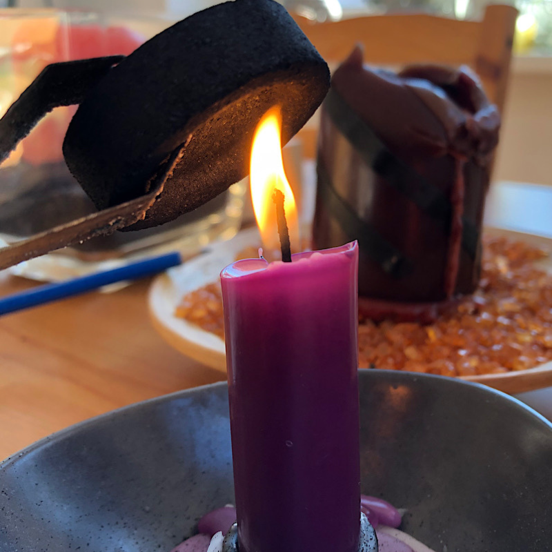 Im Vordergrund: eine brennende Kerze, an der mit einer Zange eine Räucherkohle angezündet wird. Im Hintergrund ist ein Tisch und eine weitere Kerze zu sehen. Ein Symbol für die Rauhnächte. 