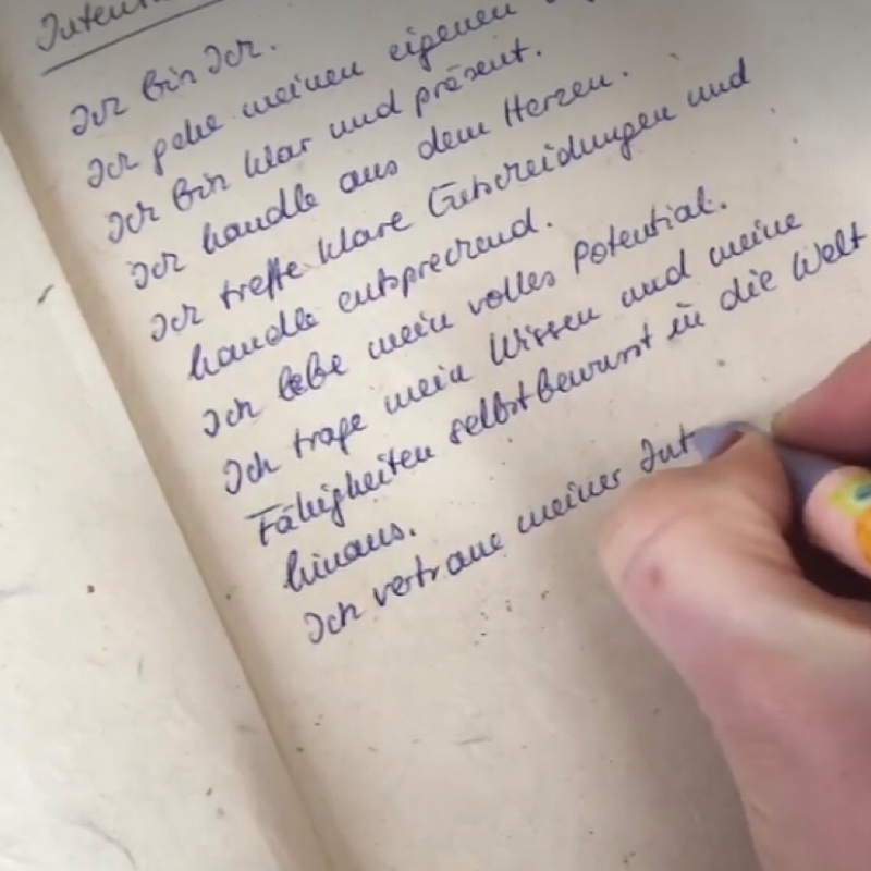 Ein Blatt Papier mit bestärkenden Sätzen: Intentionen im Rahmen der Rauhnächte. Man sieht eine Hand mit Stift, die gerade diese Sätze schreibt und einige Sätze, die bereits fertig geschrieben sind. 