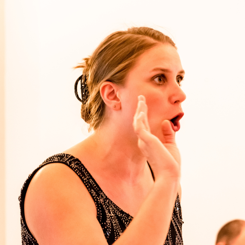 Eine Frau, die von der Seite aufgenommen ist im Portrait. Singend oder sprechend mit sehr ausdrucksstarkem Gesicht. Das Gesicht zeigt Überraschung. Die rechte Hand ist oben neben dem Gesicht und unterstützt den Ausdruck. 