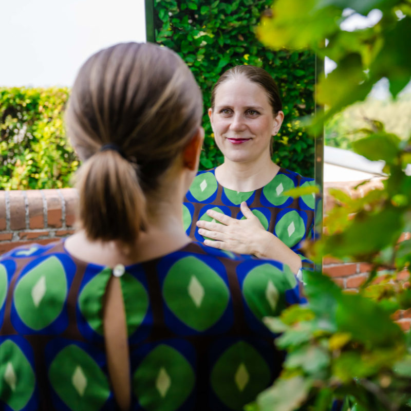 Eine Frau in einem grün-blau gemusterten Kleid steht vor einem Spiegel mitten in der Natur und hat die rechte Hand auf die Brust gelegt. Links ist eine Backstein-Mauer zu sehen. Das Foto ist von hinten aufgenommen, sodass man die Frau von hinten sieht und gleichzeitig ihr Spiegelbild von vorn. Ein Symbol dafür, wie The Turn - klar & kraftvoll das Jahr beenden durch den Kontakt mit sich selbst Klarheit und Lösung bringt.