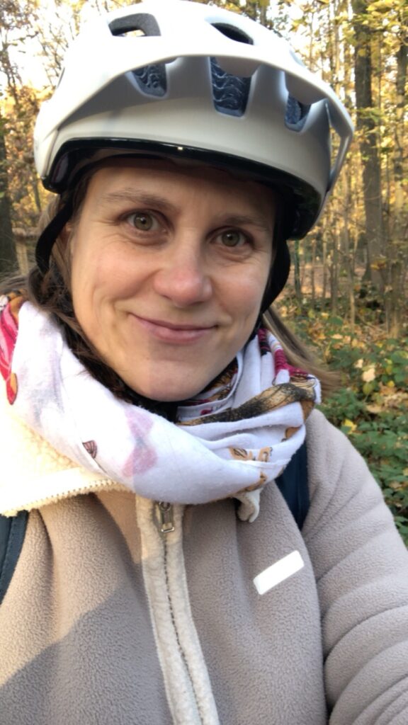 Generose mit beiger Jacke, Schal und einem Fahrradhelm auf dem Kopf lächelt in die Kamera. Im Hintergrund Wald.