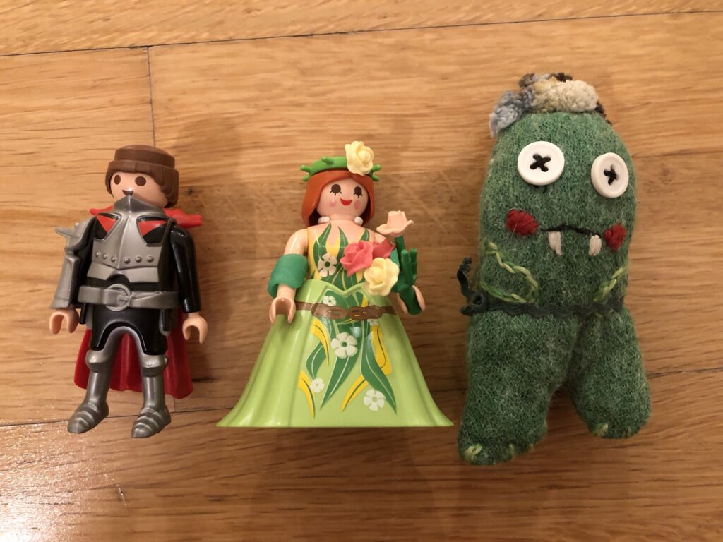 Es sind drei Figuren auf dem Bild zu sehen. Links eine Art Ritter als Playmobilfigur, in der Mitte eine Prinzessin in einem grünen Kleid, auch eine Playmobilfigur. Und rechts ein grünes kleines Monster aus Filz. Es sind Figuren, die innerhalb einer meiner IFS-Sitzungen zum Einsatz kamen.