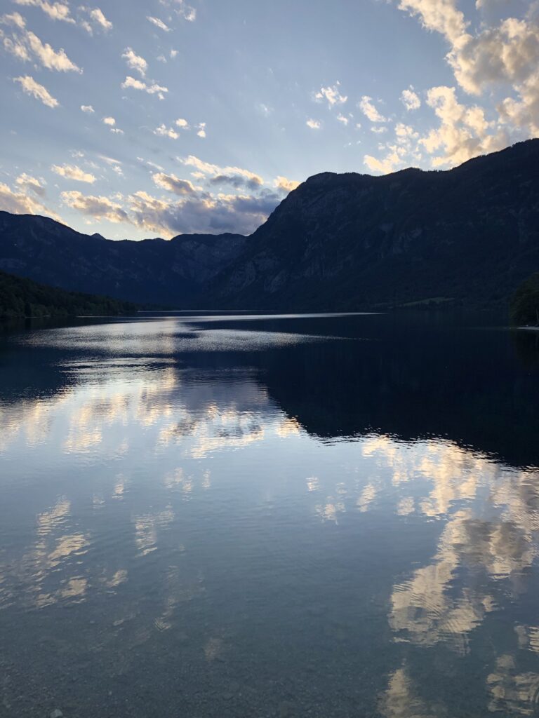 Aufnahme von einem See bei Dämmerung. Im Hintergrund ist eine Bergkette zu sehen und die Wolken und der Himmel spiegeln sich im Wasser.