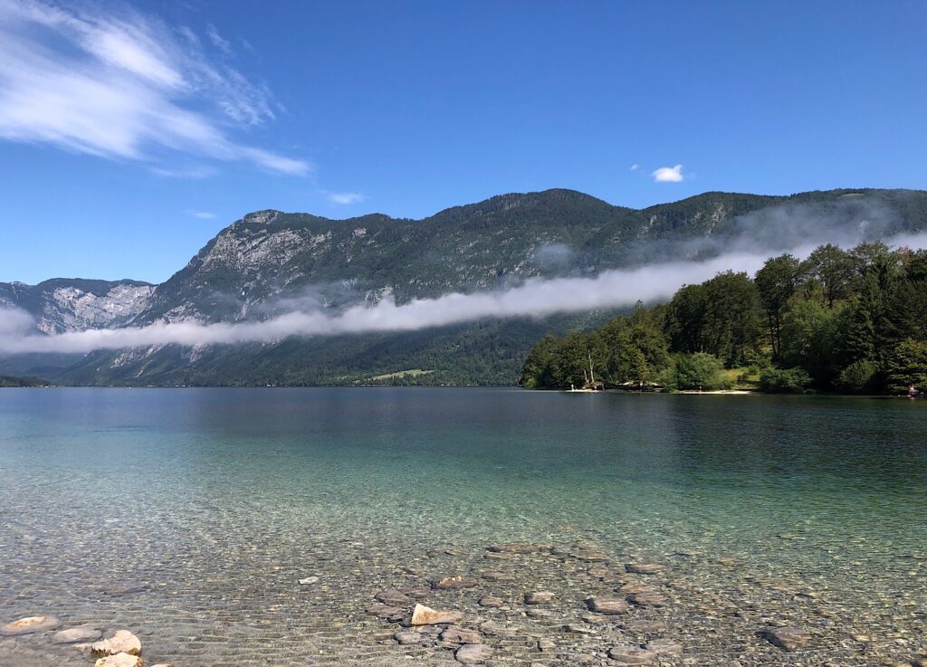 Ein See im Vordergrund, eine Bergkette im Hintergrund durchzogen von weißen Wolken, blauer Himmel und ein paar weiße Wolken am Himmel.