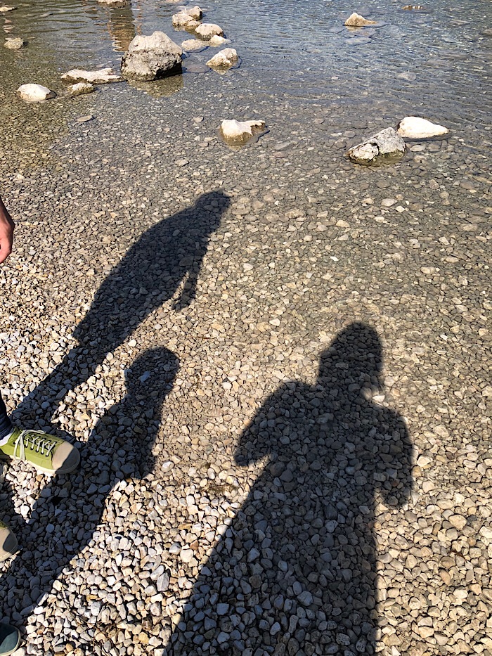 Die Schatten von drei Menschen. Zwei Erwachsenen und einem Kind. Die Schatten sind zu sehen auf kleinen Steinen, die mit Wasser bedeckt sind.