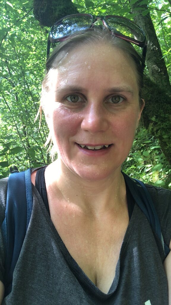 Generose mit Sonnenbrille auf dem Kopf völlig verschwitzt bei einer Wanderung. Im Hintergrund ist grüne Landschaft zu sehen.