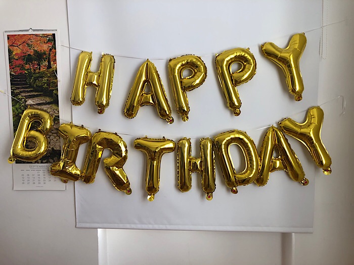 Ein goldenes Luftballon-Happy-Birthday hängt an einer weißen Wand. Daneben ein Wandkalender.