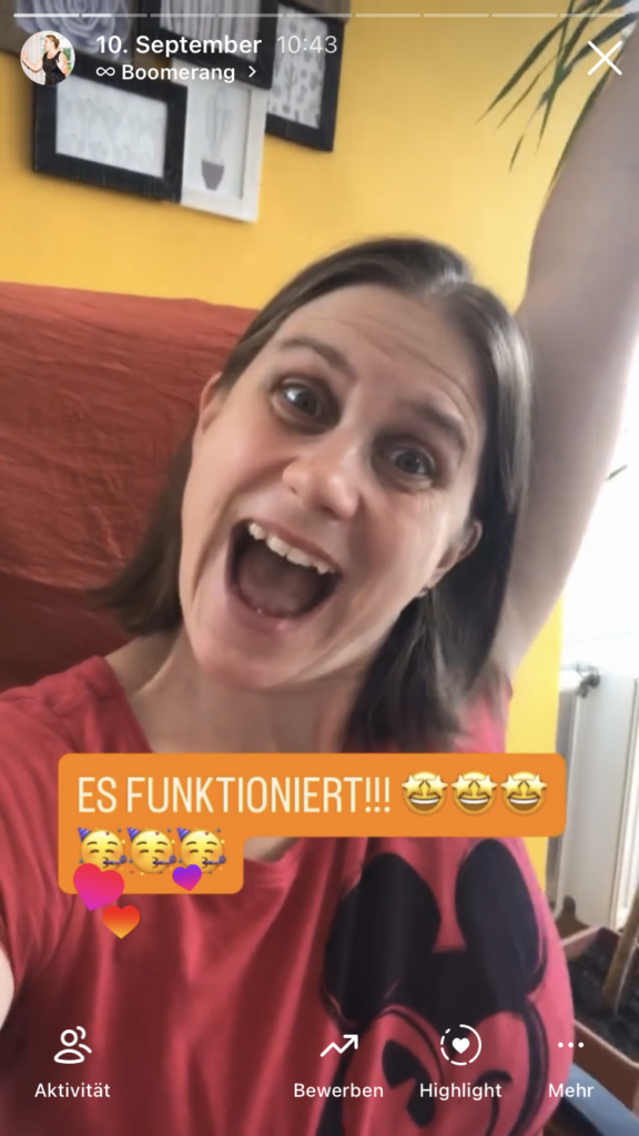 Generose jubelt mit hochstrecktem Arm in die Kamera. Im Hintergrund eine gelbe Wand mit einem Bilderrahmen und ein orangefarbener Stuhl. Auf dem Bild steht: "ES FUNKTIONIERT!"