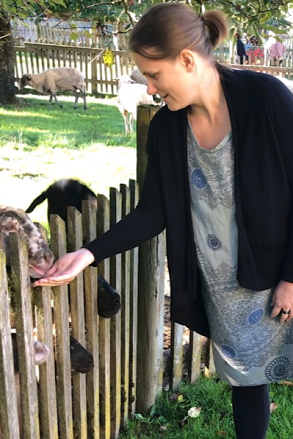 Generose in einem grünen Kleid mit einer schwarzen Weste drüber füttert gerade zwei Schafe, die hinter einem braunen Zaun sind.
