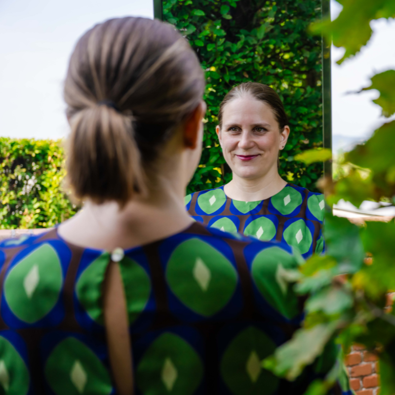 Generose blickt freundlich in einen Spiegel. Das Foto ist von hinten aufgenommen, so dass man im vorderen Teil des Bildes ihren Hinterkopf mit einem Pferdeschwanz sieht und dann im hinteren Teil des Bildes ihr Gesicht und ihren Oberkörper im Spiegel. Sie trägt ein blau grün gemustertes Kleid und am Rand des Bildes sind Hecken und Blätter zu sehen. 