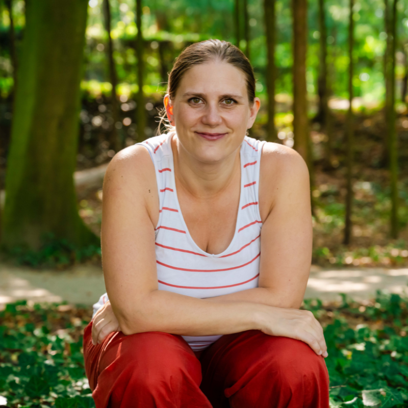 Generose kniet in der Hocke mit auf den Beinen überschlagenen Armen mitten im Wald. Im Hintergrund etwas Gras und Baumstämme. Sie trägt ein weißes Shirt mit orangen Streifen und eine orange Hose und schaut direkt frontal freundlich in die Kamera.