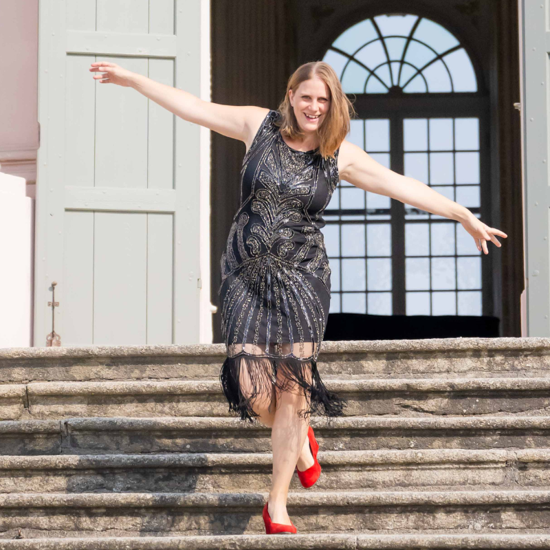 Generose steht im schwarzen Kleid mit Glitzerpailletten und roten Schuhen auf einer steinernen Treppe und hat die Arme ausgebreitet. Sie lacht und geht gerade spielerisch die Treppe runter. Im Hintergrund ist eine offene Tür und ein barockes Fenster mit einem Gitter zu sehen. Ein Sinnbild für Selbstbewusstsein ohne People Pleasing.