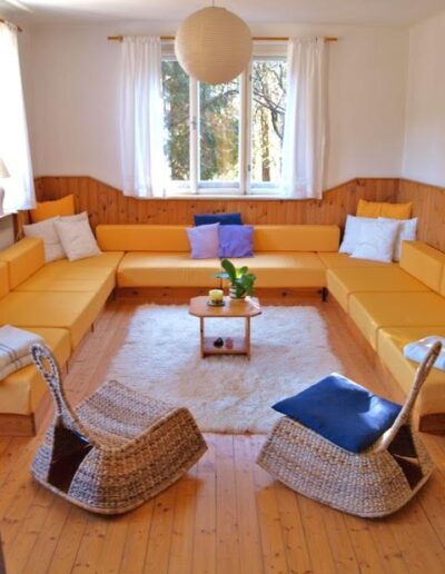 Ein Zimmer von der Tür aus in Richtung Fenster fotografiert mit einer U-förmigen Sitzbank in gelb und zwei gemütlichen Korbsesseln davor. Teppich auf dem Boden und einem kleinen Tischchen in der Mitte.