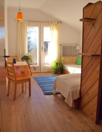 Von der Tür aus in ein Zimmer hineinfotografiert in Richtung Balkontür. Es sind zwei Betten, ein Stuhl und ein Tisch zu sehen. Neben dem Fenster steht eine Pflanze und auf dem Boden liegt ein blau gestreifter Teppich.