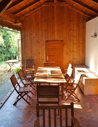 Foto von einer überdachten Holzterrasse mit Holztisch und Holzstühlen, die Sonne scheint gerade und im Hintergrund sind Bäume und Grünfläche zu sehen.