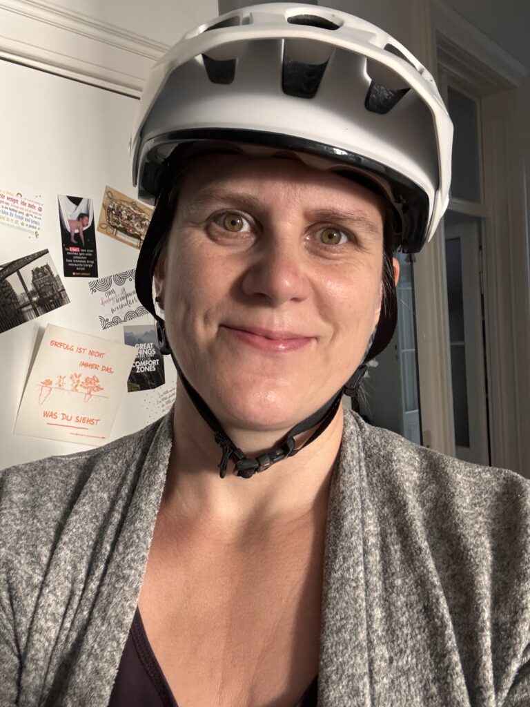 Ein Selfie von Generose mit einer grauen Weste und einem weißen Fahrradhelm. Im Hintergrund eine Tür mit Postkarten. 