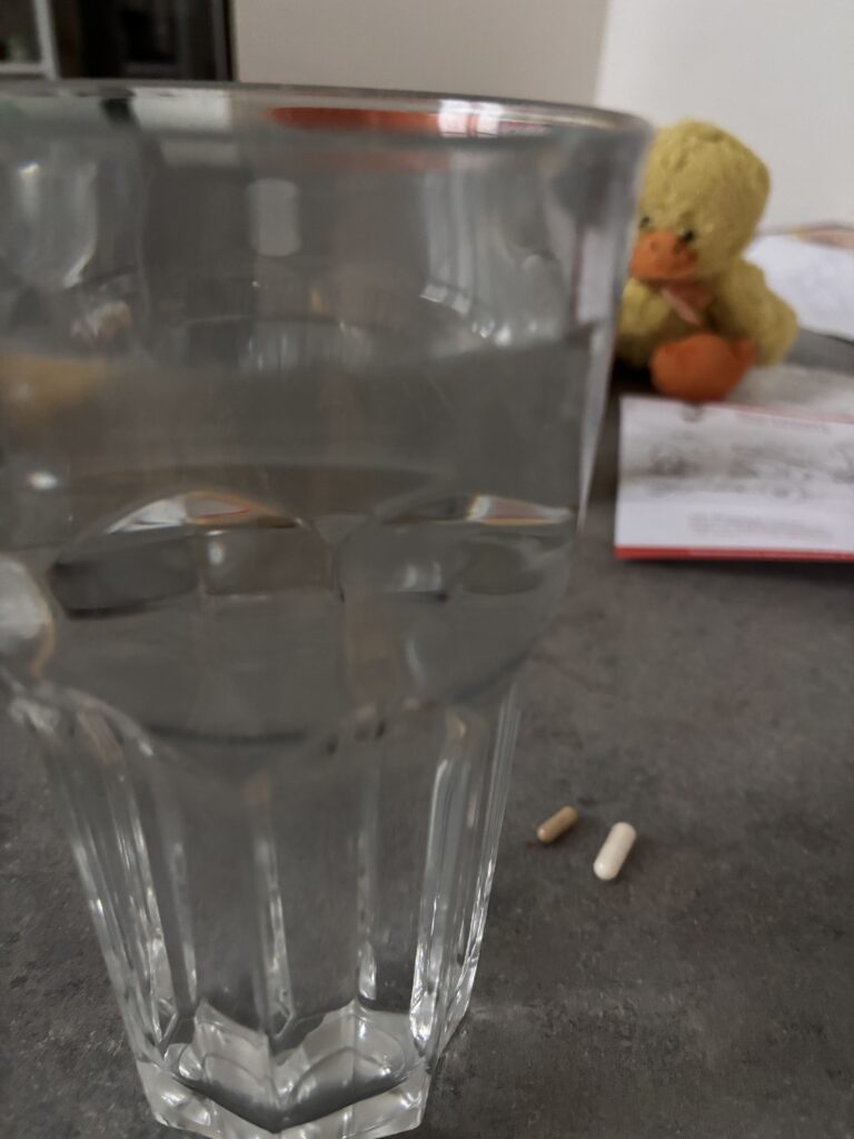 Im Vordergrund ein großes Glas mit Wasser gefüllt, daneben zwei kleine Tabletten. Im Hintergrund der Küchentisch und eine Stofftier-Ente.